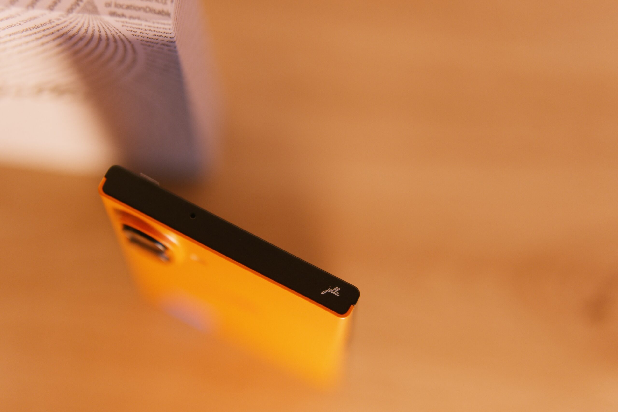 Das Jolla Phone von oben. Rechts sieht man ein kleines jolla Logo. Im Hintergrund die weiße Verpackung mit jolla Schriftzug umschlungen von einer Buchstabenspirale (Programmiercode)
