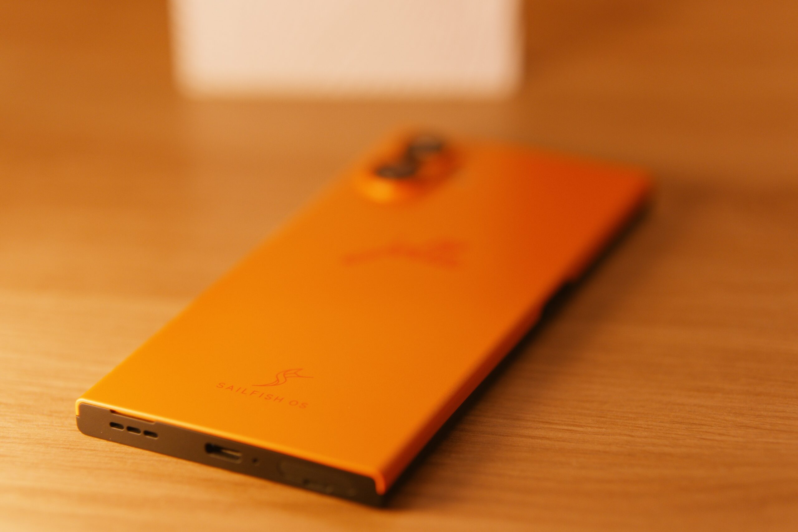 Das jolla Phone liegt am Bauch. Die Rückseite (Orange). Links oben unscharf Kamerabuckel + LED, mittig jolla Schriftzug. Unteres Drittel Sailfish OS Schriftzug mit Logo (S wie ein Segeltuch) Auf der Unterseite sieht man v.l.n.r Lautsprecher, USB-C, Mikrofon, SIM / MSD Tray. 