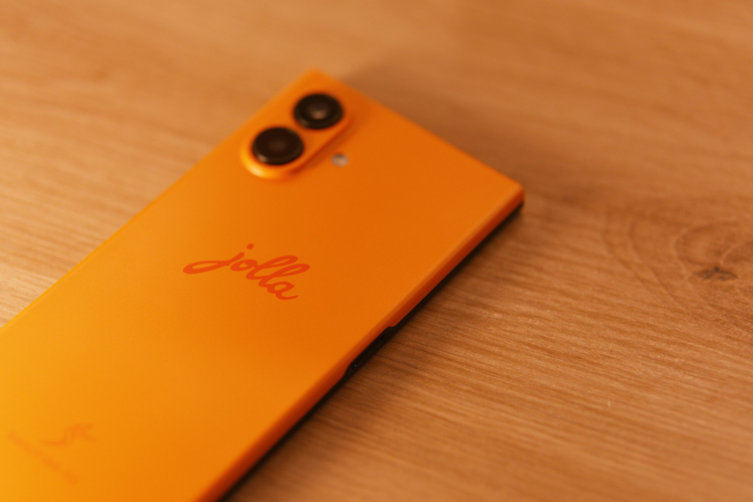 Das jolla Phone liegt am Bauch. Die Rückseite (Orange). Links oben unscharf Kamerabuckel + LED, mittig jolla Schriftzug. Unteres Drittel Sailfish OS Schriftzug mit Logo (S wie ein Segeltuch).