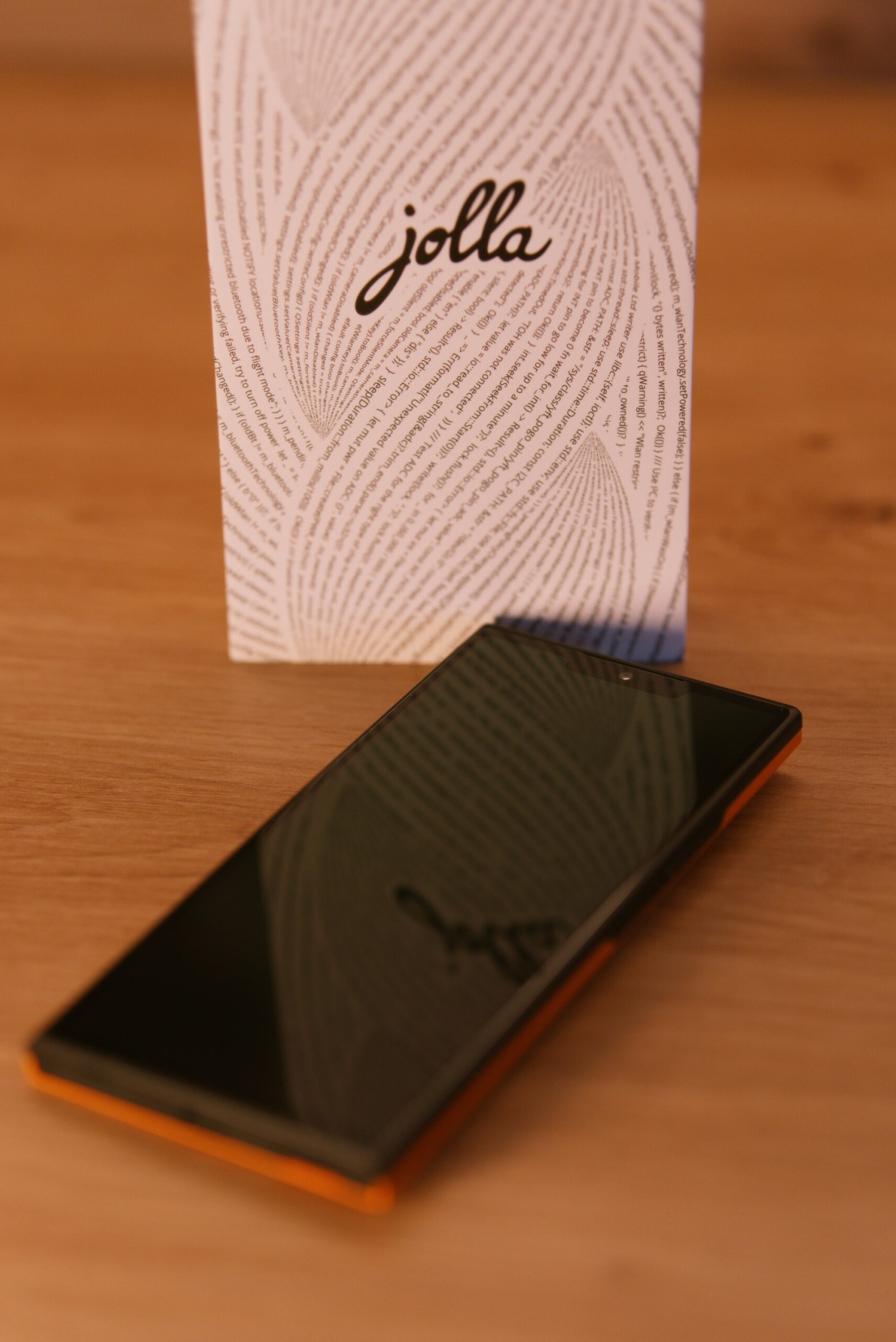 Das jolla Phone von vorne. Man sieht das Display im ausgeschalteten Zustand. Mittig eine "Notch" mit Selfie Kamera. Im Hintergrund die weiße Verpackung mit jolla Schriftzug umschlungen von einer Buchstabenspirale (Programmiercode)
