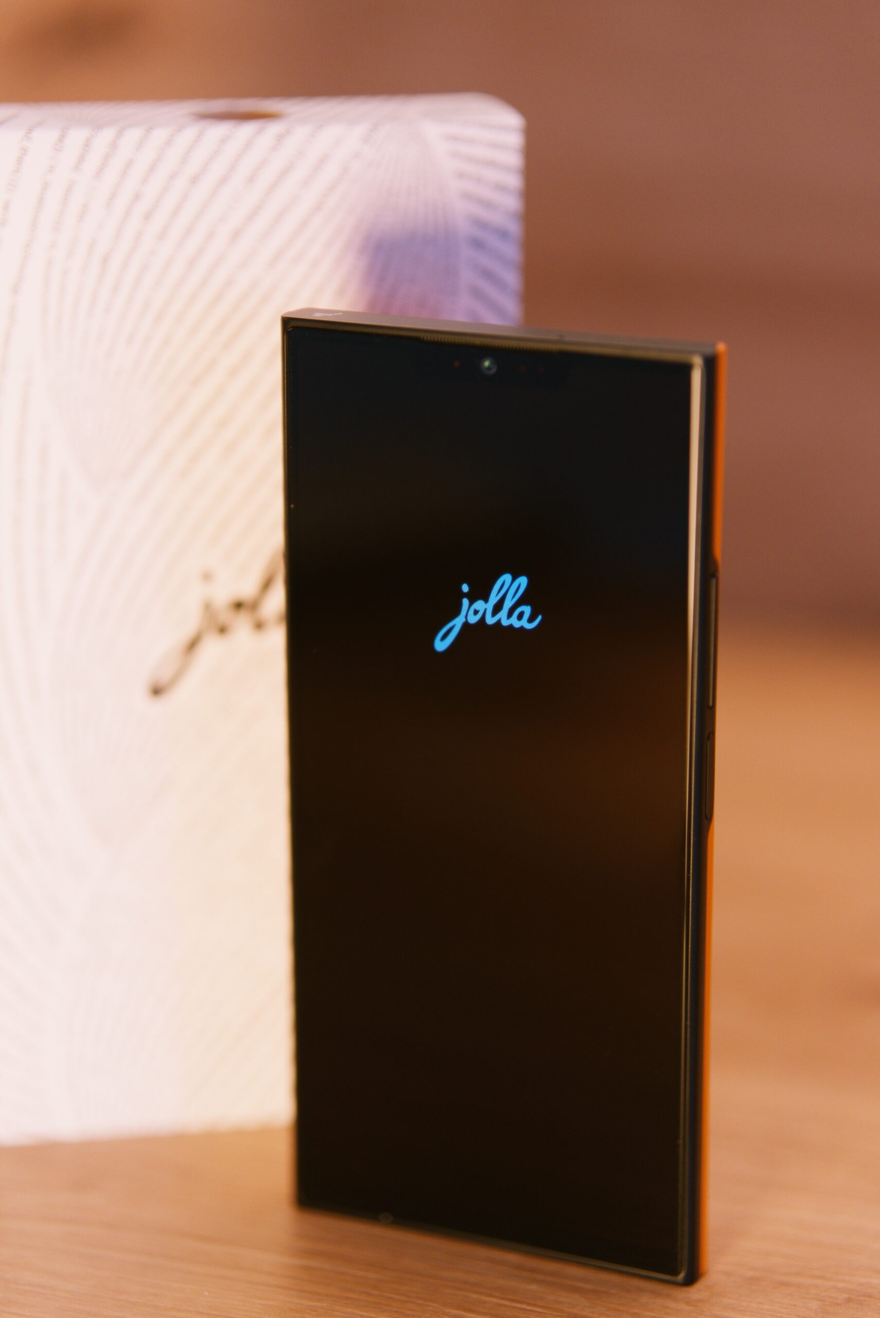 Das jolla Phone von vorne. Man sieht das Display eingeschaltet mit jolla Schriftzug. Mittig oben eine "Notch" mit Selfie Kamera. Im Hintergrund die weiße Verpackung mit jolla Schriftzug umschlungen von einer Buchstabenspirale (Programmiercode)