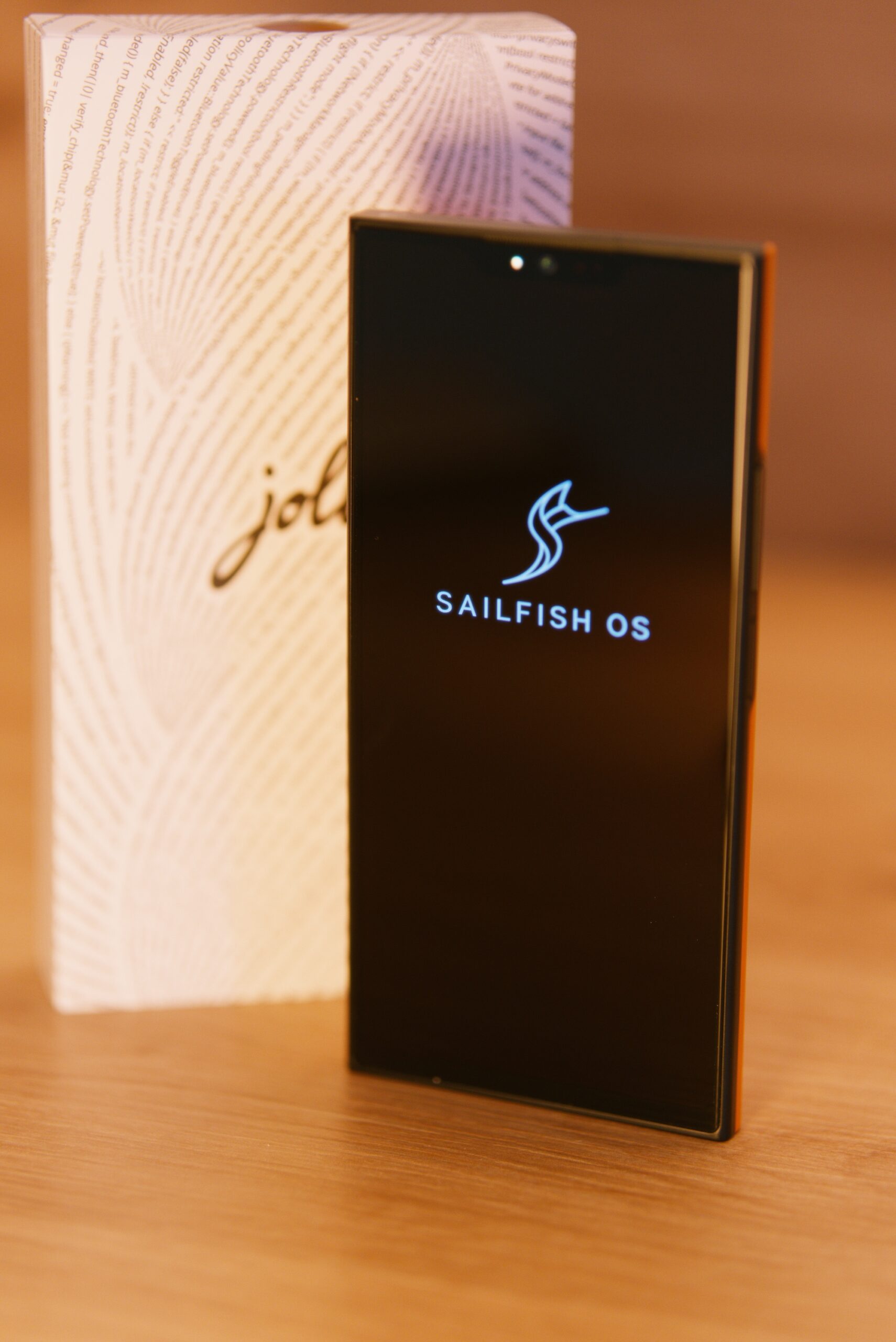 Das jolla Phone von vorne. Man sieht das Display eingeschaltet mit Sailfish OS Schriftzug und einen Logo (S wie ein Segeltuch). Mittig oben eine "Notch" mit Selfie Kamera. Im Hintergrund die weiße Verpackung mit jolla Schriftzug umschlungen von einer Buchstabenspirale (Programmiercode)