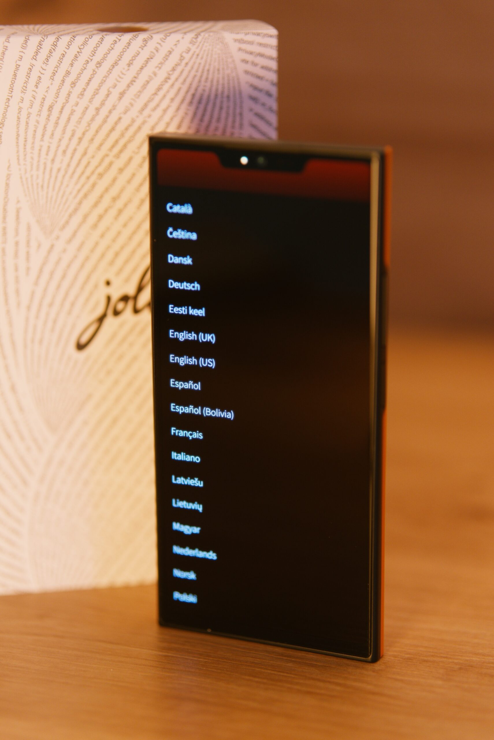 Das jolla Phone von vorne. Man sieht das Display eingeschaltet mit einen Auswahlbildschirm verschiedenster Sprachen. Mittig oben eine "Notch" mit Selfie Kamera. Im Hintergrund die weiße Verpackung mit jolla Schriftzug umschlungen von einer Buchstabenspirale (Programmiercode)