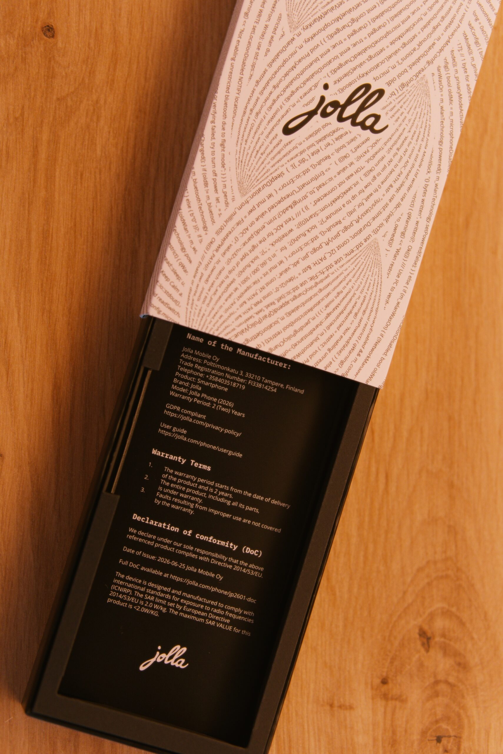 Zettelchen in der Schalter mit folgenden Text.
Name of the Manufacturer:
Jolla Mobile Oy
Address: Polttimonkatu 3, 33210 Tampere, Finland
Trade Registration Number: FI33814254
Telephone: +358403518719
Product: Smartphone
Brand: Jolla
Model: Jolla Phone (2026)
Warranty Period: 2 (Two Years
GDPR compliant
https://jolla.com/privacy-policy/
User guide
https://jolla.com/phone/userguide
Warranty Terms
1. The warranty period starts from the date of delivery of the product and is 2 years.
The entire product, including all its parts, is under warranty.
3. Faults resulting from improper use are not covered by the warranty.
Declaration of conformity (DoC)
We declare under our sole responsibility that the above referenced product complies with Directive 2014/53/EU.
Date of Issue: 2026-06-25 Jolla Mobile Oy
Full DoC available at https://jolla.com/phone/jp2601-doc
The device is designed and manufactured to comply with international standards for exposure to radio frequencies
(ICNIRP). The SAR limit set by European Directive
2014/53/EU is 2.0 W/kg. The maximum SAR VALUE for this product is <2.0W/KG.
 Im Hintergrund die weiße Verpackung mit jolla Schriftzug umschlungen von einer Buchstabenspirale (Programmiercode)