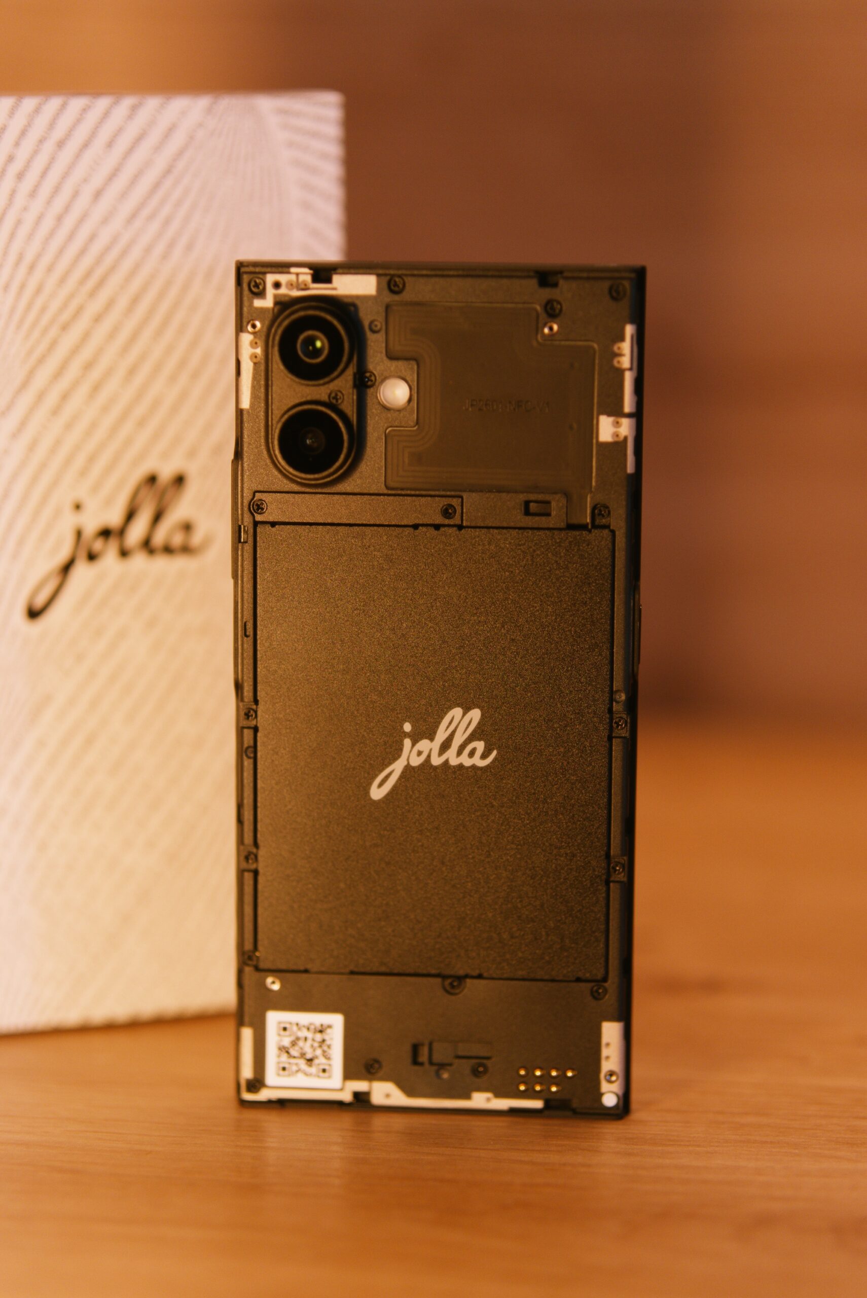 Das jolla Phone stehend ohne Cover. Man sieht dass der Akku getauscht werden kann mit Schrauben. Rechts unten Pins für The Other Half. Im Hintergrund die weiße Verpackung mit jolla Schriftzug umschlungen von einer Buchstabenspirale (Programmiercode)