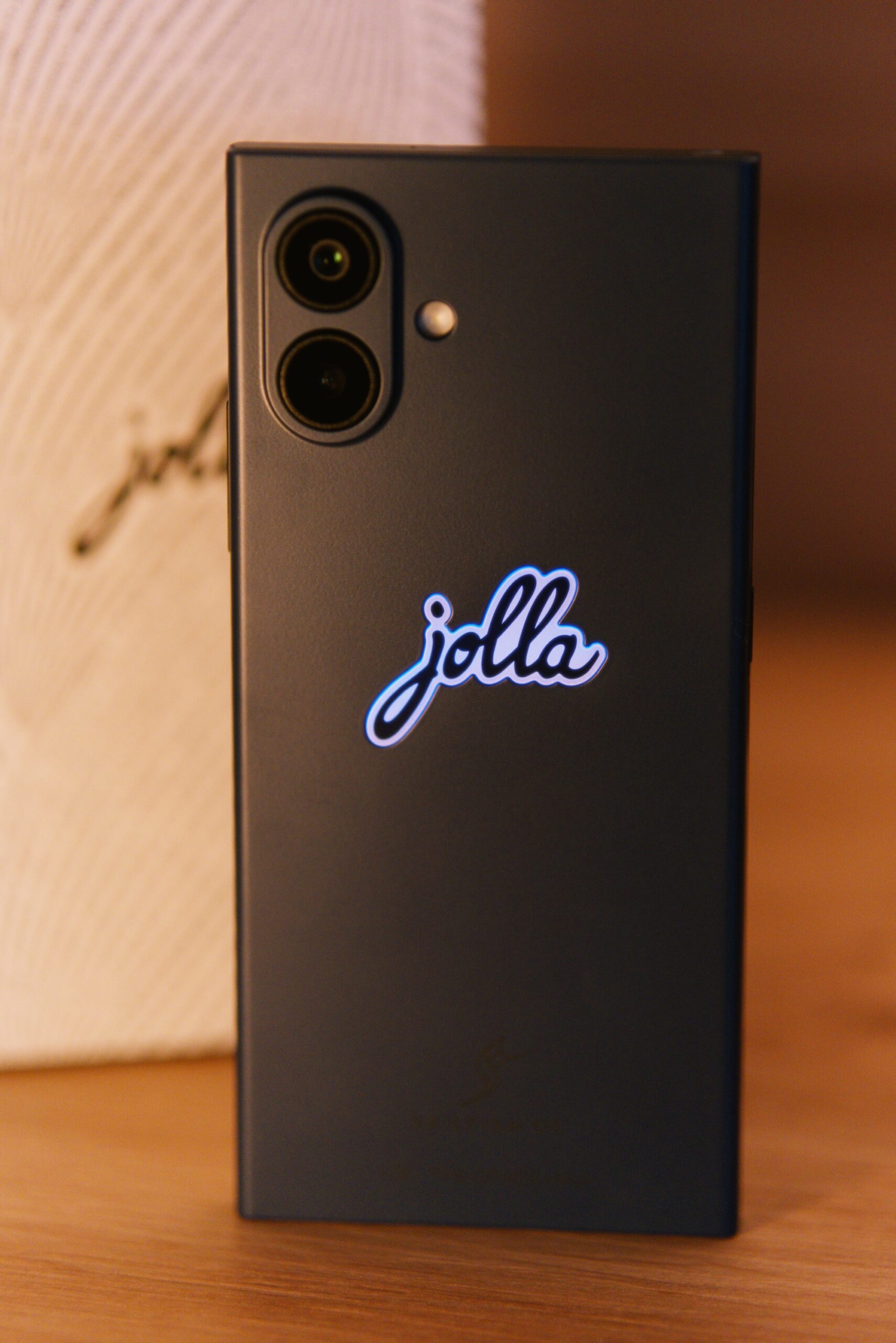 The Other Half in Inari Blue. Mittig das jolla Logo am Cover beleuchtet.  Im Hintergrund die weiße Verpackung mit jolla Schriftzug umschlungen von einer Buchstabenspirale (Programmiercode)