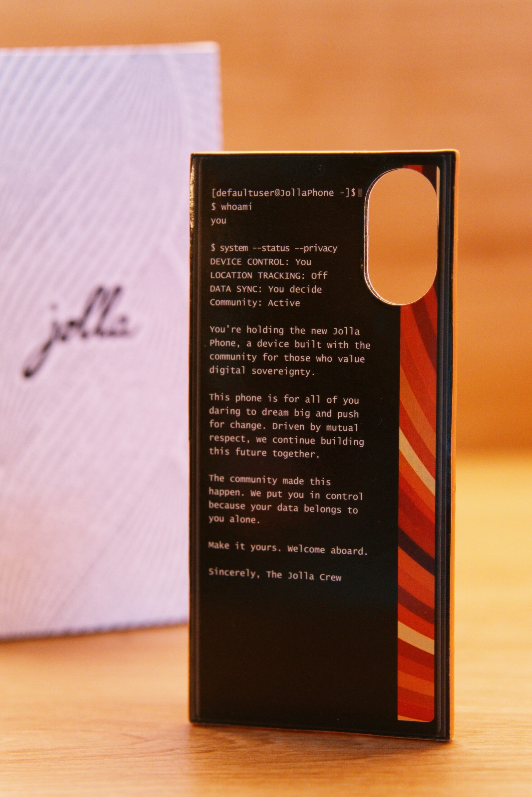 Grußkarte von der Jolla Crew. Mit folgenden Text:
[defaultuser@Jollaphone -1$
$ whoami you
$ system --status --privacy
DEVICE CONTROL: You
LOCATION TRACKING: off
DATA SYNC: You decide
Community: Active
You're holding the new Jolla . Phone, a device built with the community for those who value digital sovereignty.
This phone is for all of you daring to dream big and push for change. Driven by mutual respect, we continue building this future together.
The community made this happen. We put you in control because your data belongs to you alone.
Make it yours. welcome aboard.
Sincerely, The Jolla Crew
 Im Hintergrund die weiße Verpackung mit jolla Schriftzug umschlungen von einer Buchstabenspirale (Programmiercode)